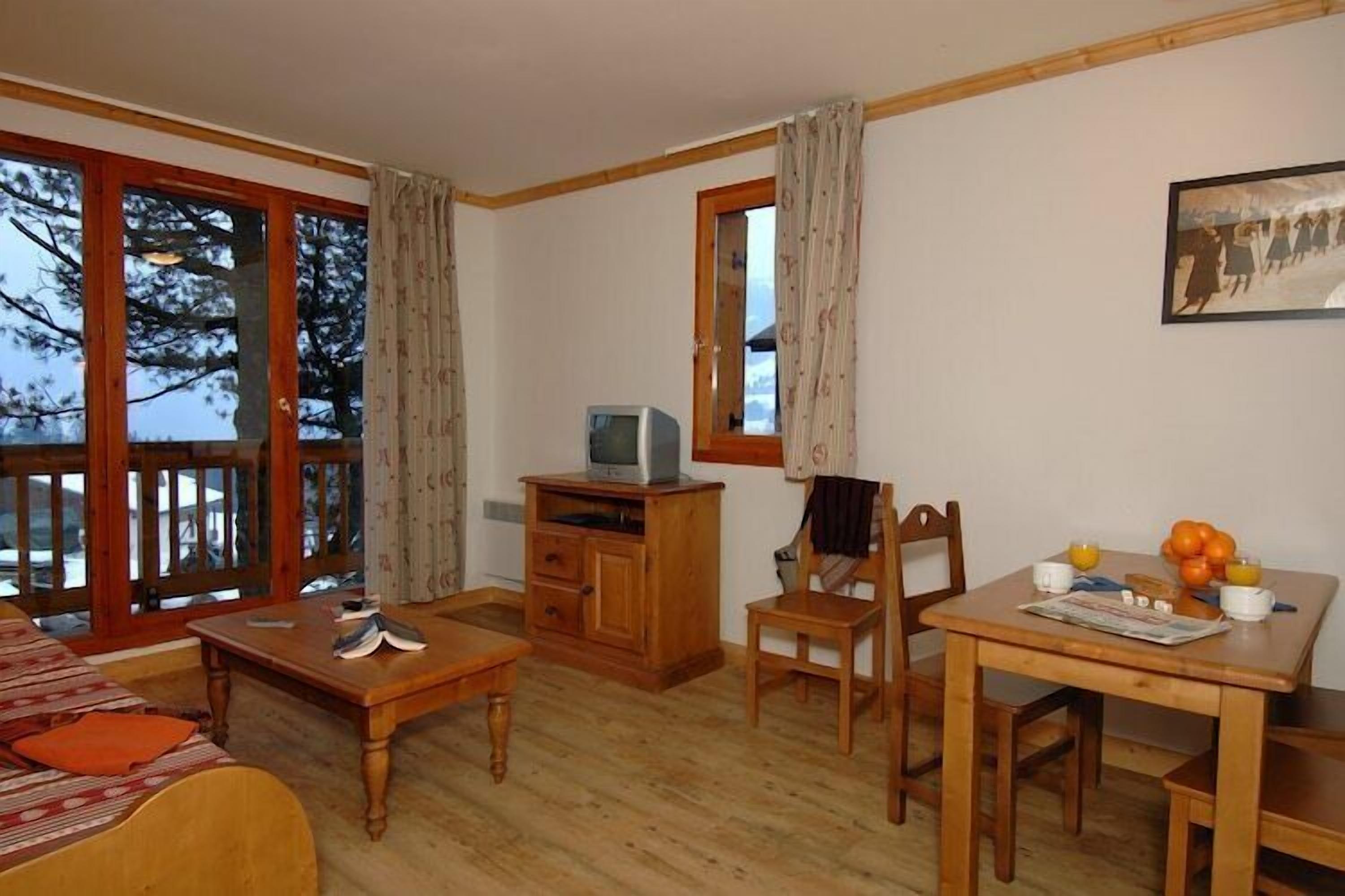Un salon meublé comprenant un canapé, une table basse, un meuble pour la télévision et une table à manger avec des chaises. Il y a une grande fenêtre avec des rideaux et un tableau accroché au mur.