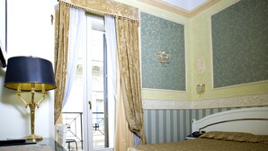 A hotel room with a bed, a desk with a lamp, a window with curtains, and a wall with decorative moldings. - Upper View Suite (Rome)