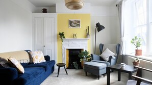 Ein Wohnzimmer mit blauem Sofa, grauem Sessel, kleinem Tisch und Kamin, dessen Wand in Gelb gehalten ist.