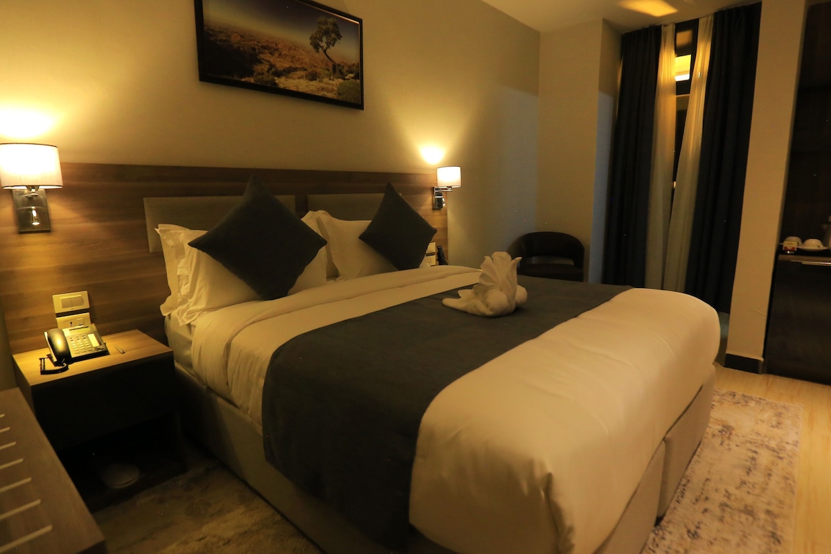 Habitación de hotel con cama, lámparas de noche, teléfono, escritorio y un cuadro en la pared.
