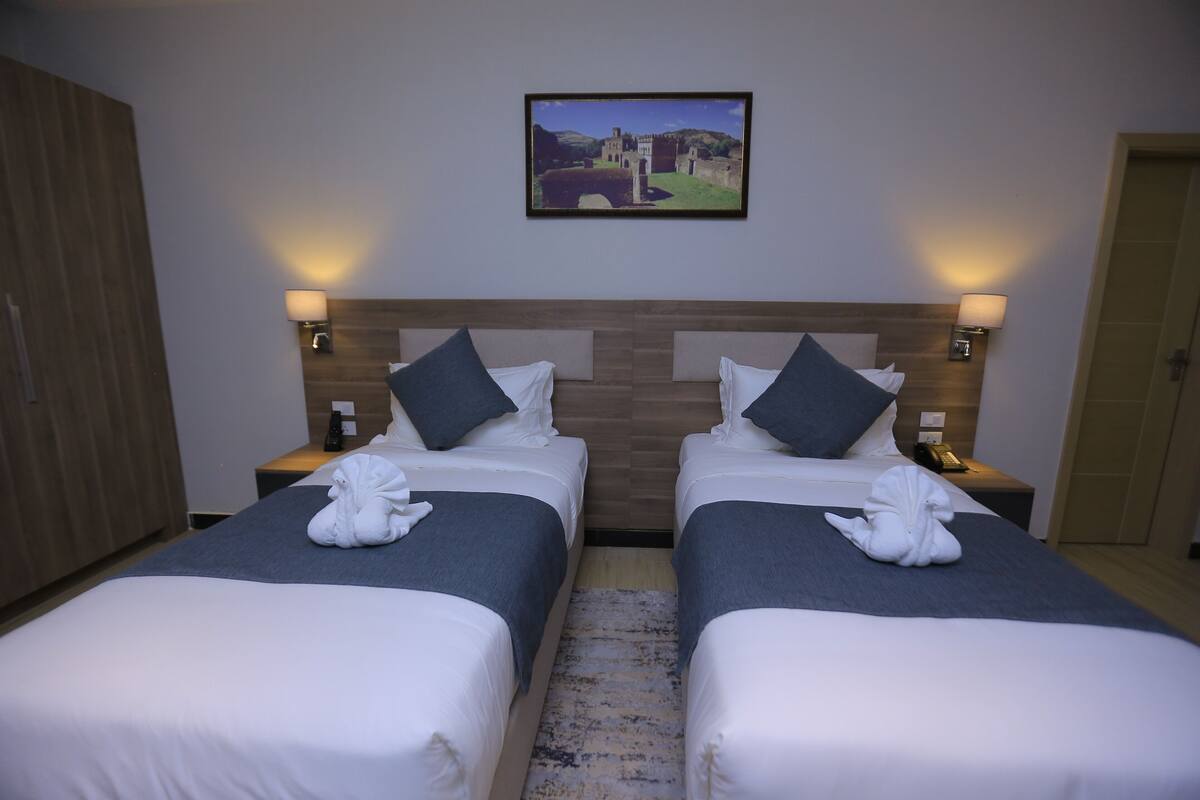 A hotel room with two beds, a wooden headboard, a framed picture on the wall, and a small table with a lamp.