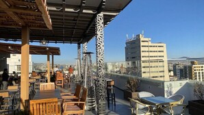 Una terraza al aire libre con mobiliario de madera y pilares decorativos, con vistas a un paisaje urbano con edificios y cielo despejado.