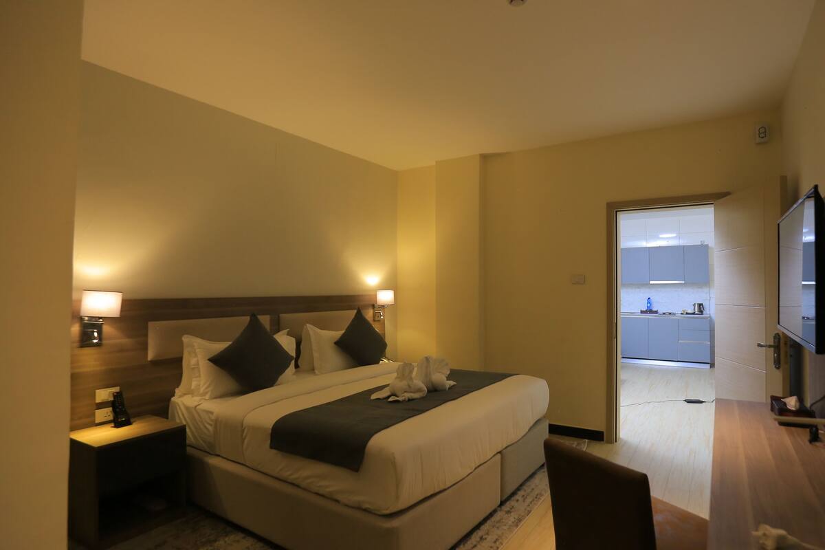 A hotel room with a bed, bedside tables, a TV, and a view of the kitchen.