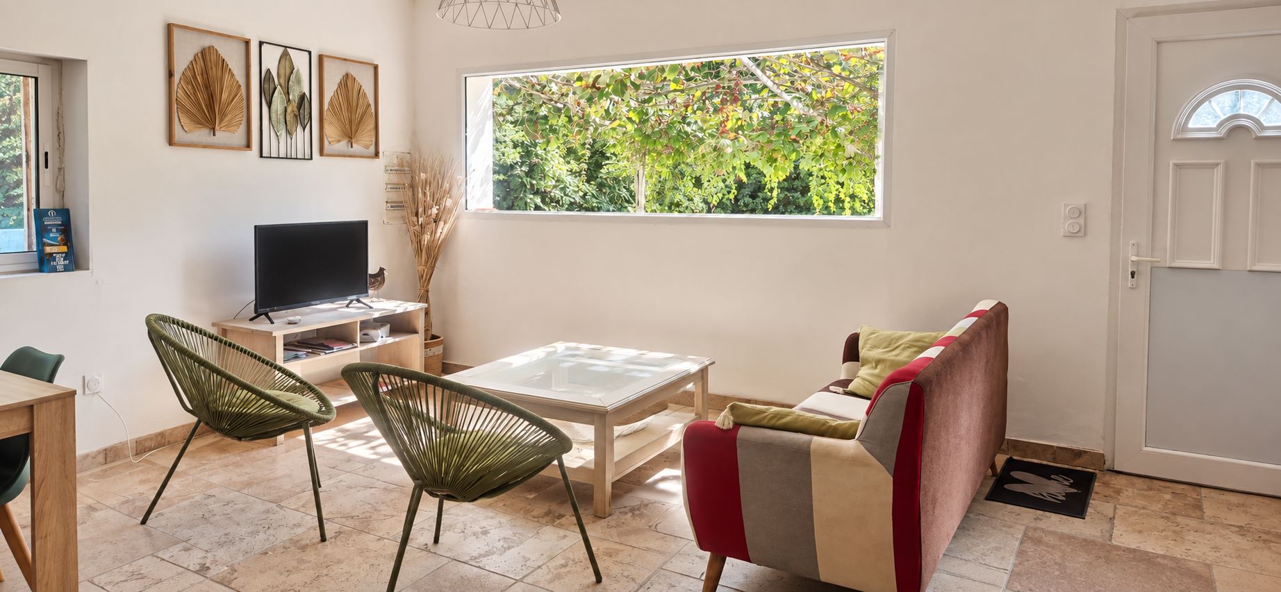A living room with a TV, two chairs, a sofa, a coffee table, and a window with a view of greenery.