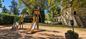 Un terrain de jeux avec un toboggan et des structures d’escalade, entouré d’arbres et bordé d’un bâtiment en pierre.