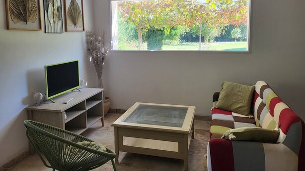 Een woonkamer met een televisie, een salontafel met glazen blad, een kleurrijke bank en een groot raam met uitzicht op bomen.
