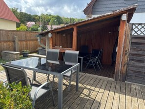 En udendørs patio med et glasbord og stole, et trædæk og et overdækket siddeområde.
