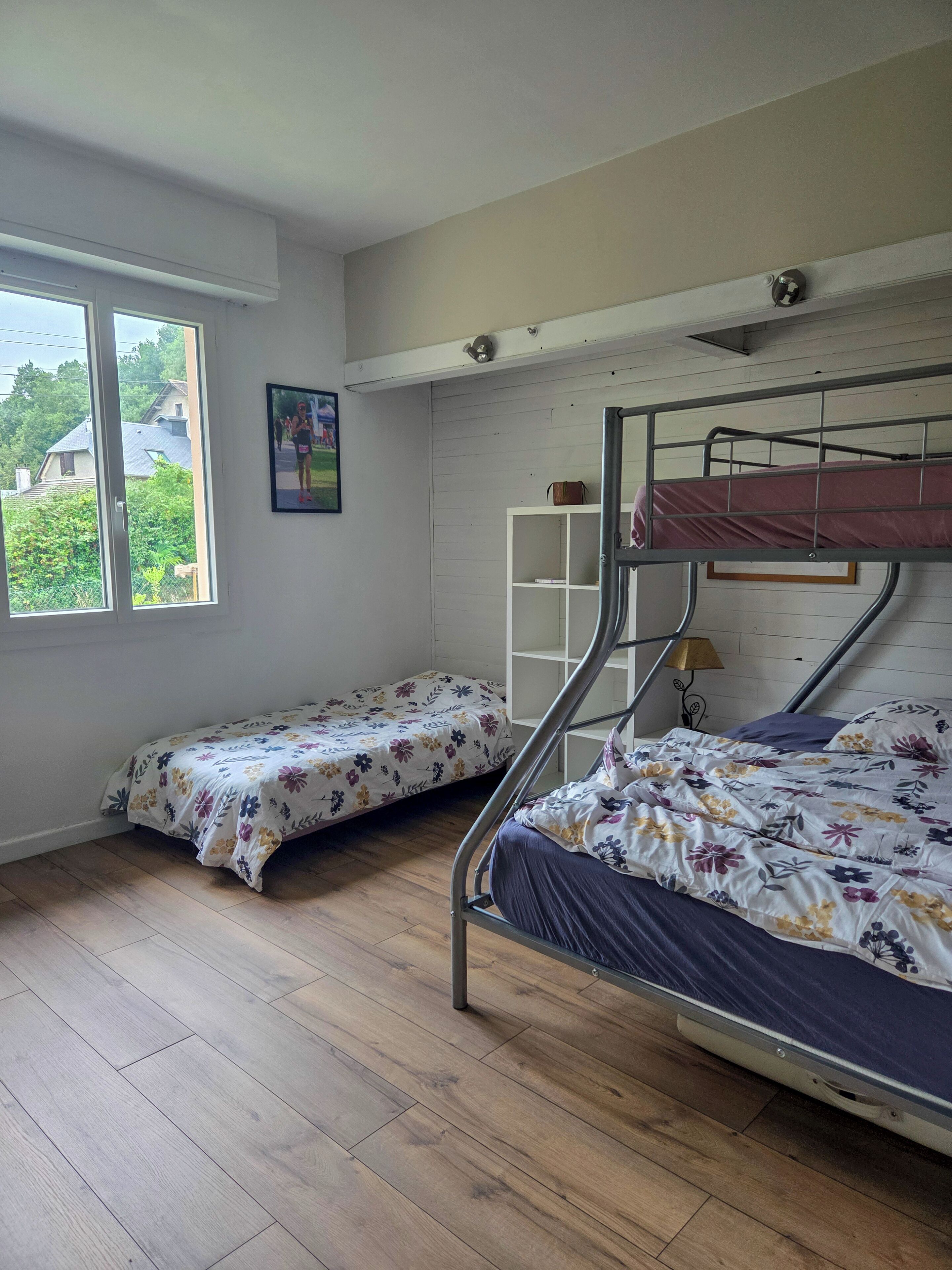 A dormitory room with two bunk beds, wooden flooring, a white wall, a window with a view of houses, and a small shelf with books.