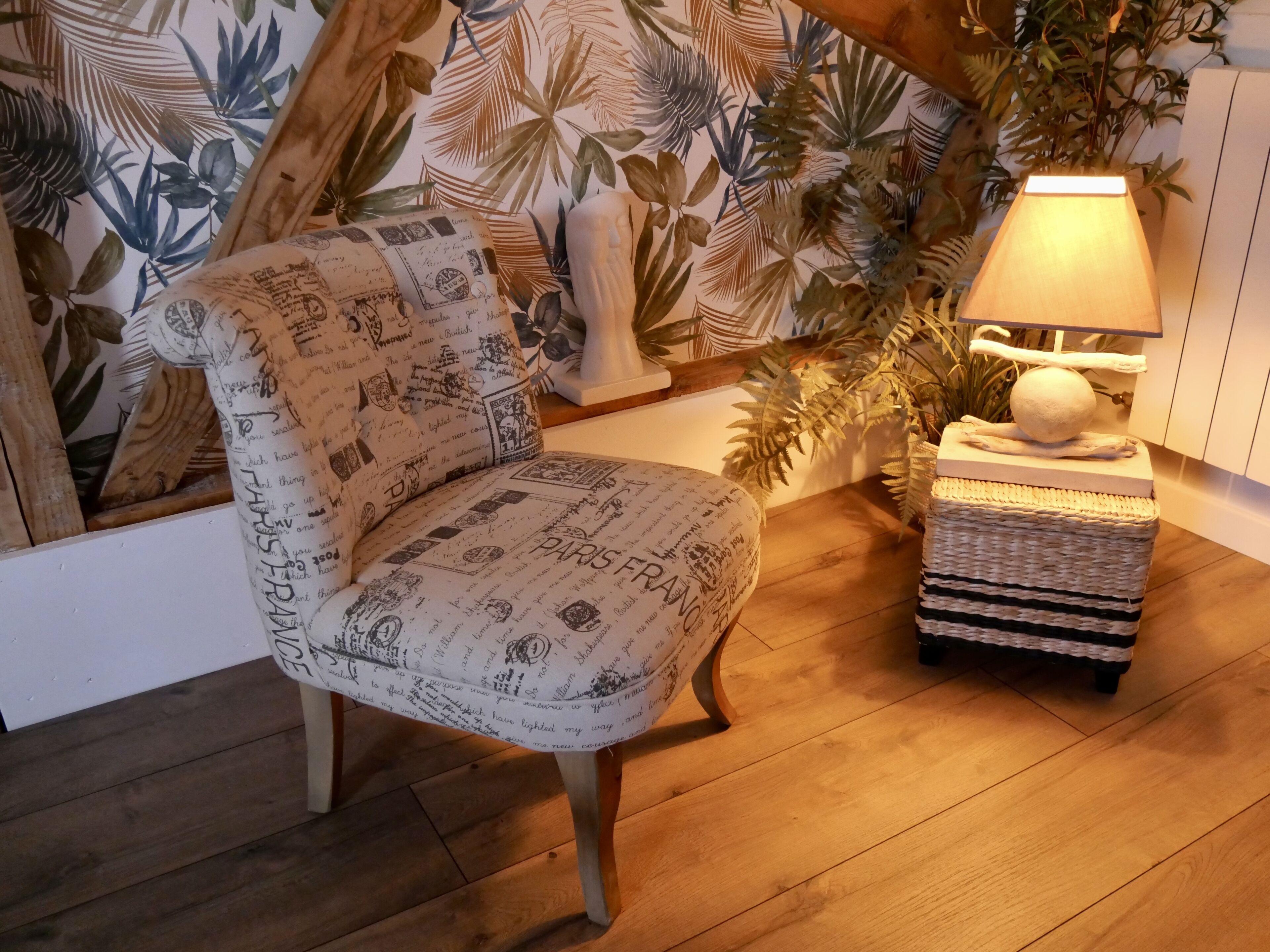 A chair with a patterned fabric, a small table with a lamp, and a wall with a tropical leaf design.