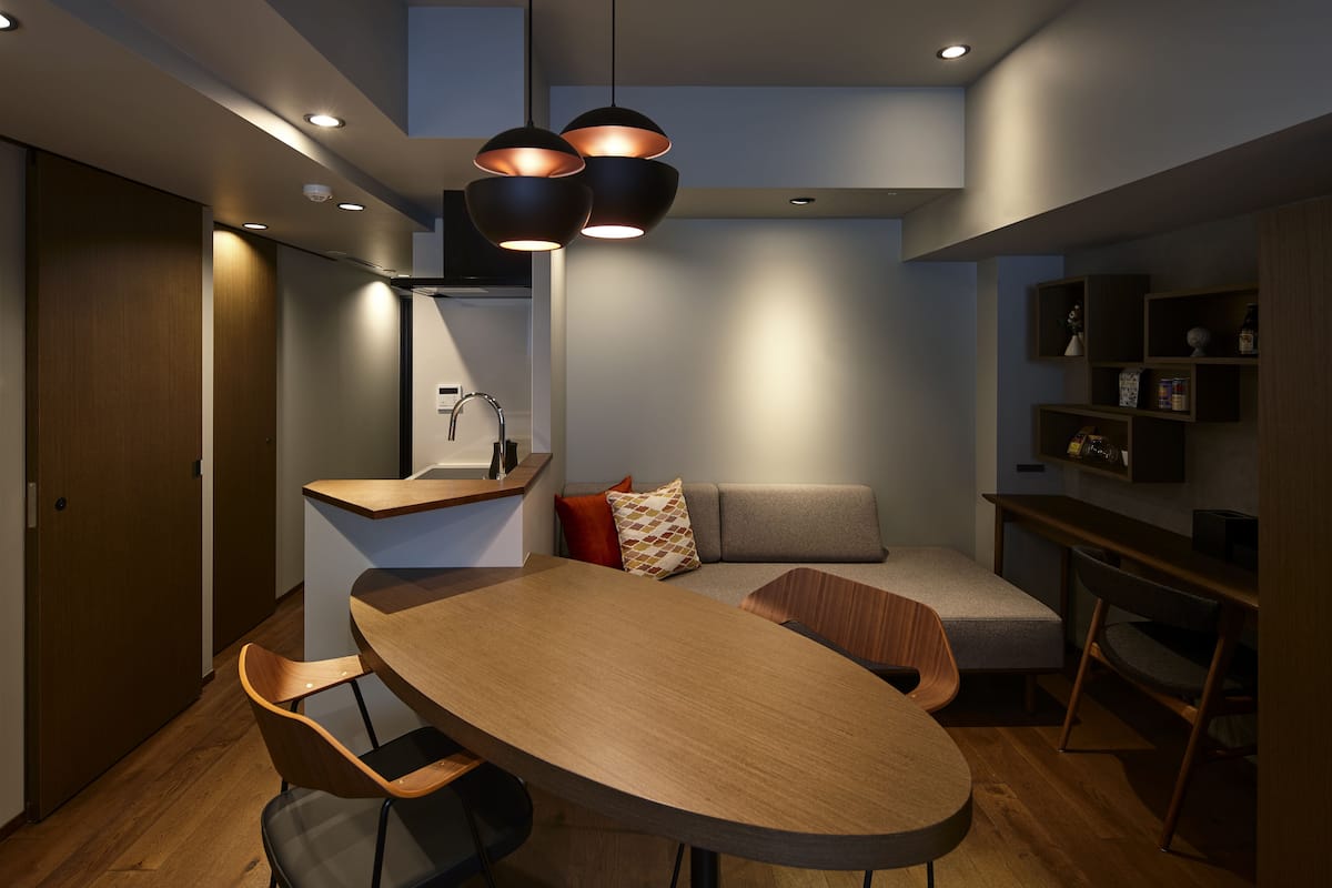 A modern interior with a wooden dining table, chairs, a sofa, and a built-in shelf with decorative items.
