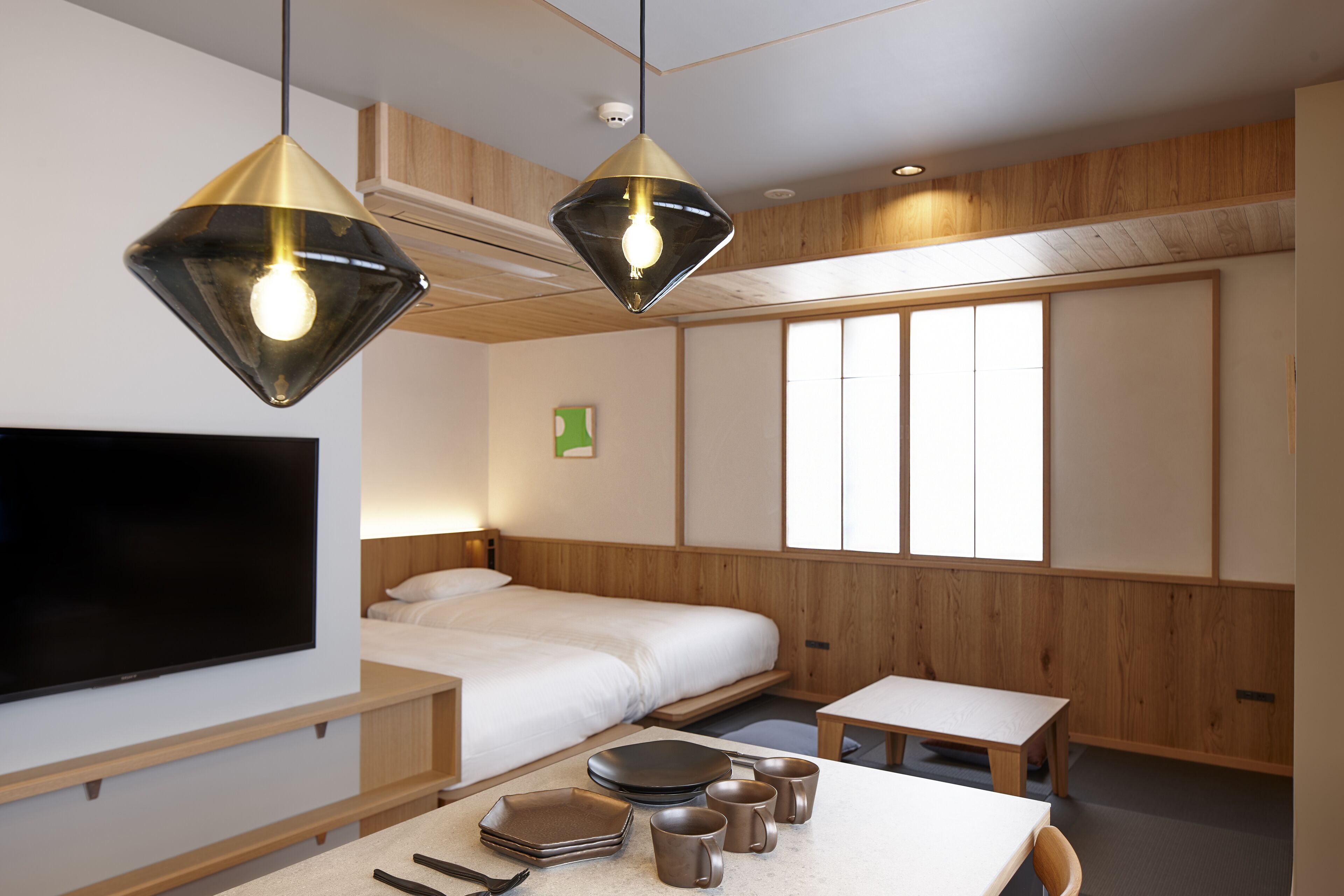 A modern hotel room with a bed, a TV, a dining table with plates and cups, and two pendant lights.