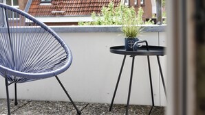 Ein Balkon mit blauem Stuhl, einem kleinen Tisch mit Topfpflanze und Blick auf ein Gebäude mit roten Ziegeldächern im Hintergrund.
