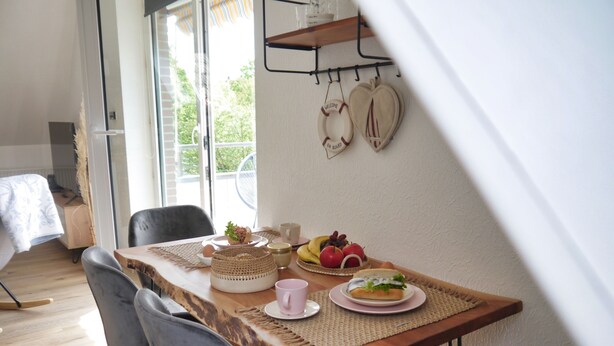 Ein Essbereich mit einem Holztisch, zwei grauen Stühlen, einer gewebten Tischmatte, einem Teller mit Obst, einem Sandwich und einem Schild an der Wand mit der Aufschrift 'Welcome on Board'.