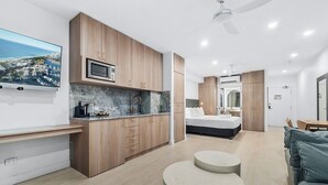 A modern kitchen with wooden cabinets, a built-in microwave, and a marble countertop. There is a bed with a white mattress and a small seating area with a round table.