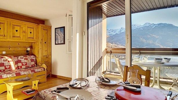Una habitación acogedora con un sofá de madera, una mesa de comedor dispuesta para dos personas y vistas a montañas nevadas a través de amplios ventanales.
