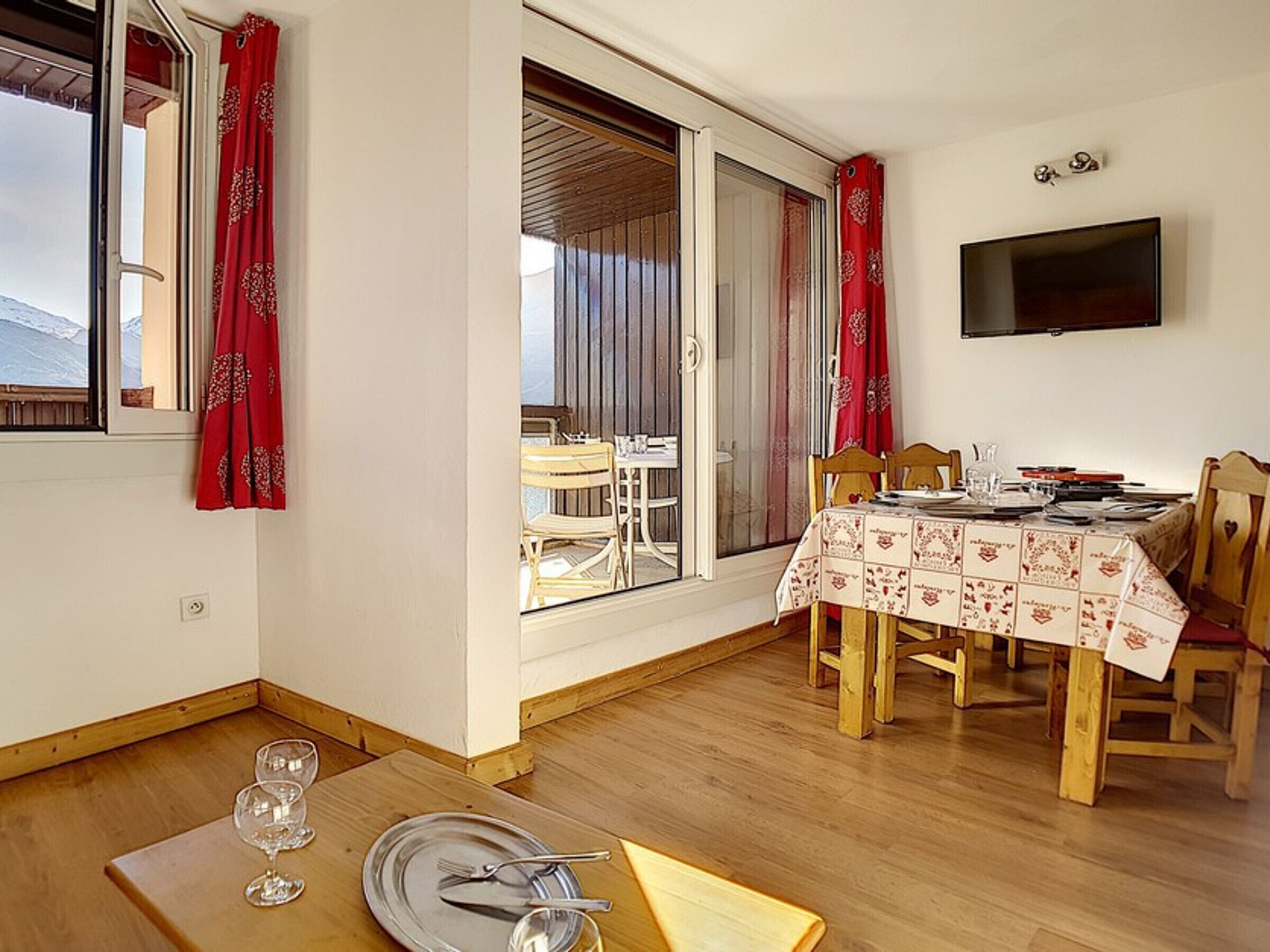 A dining area with a table set for two, a television mounted on the wall, and a view of the outdoors through a sliding glass door.