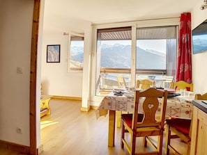 Un coin repas avec une table dressée pour quatre personnes, des chaises en bois, une vue sur les montagnes par une grande fenêtre et un rideau rouge sur le côté droit.