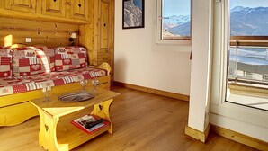 Una habitación acogedora con paredes de madera, un sofá estampado, una pequeña mesa de madera con una revista y una ventana con vistas a la montaña.