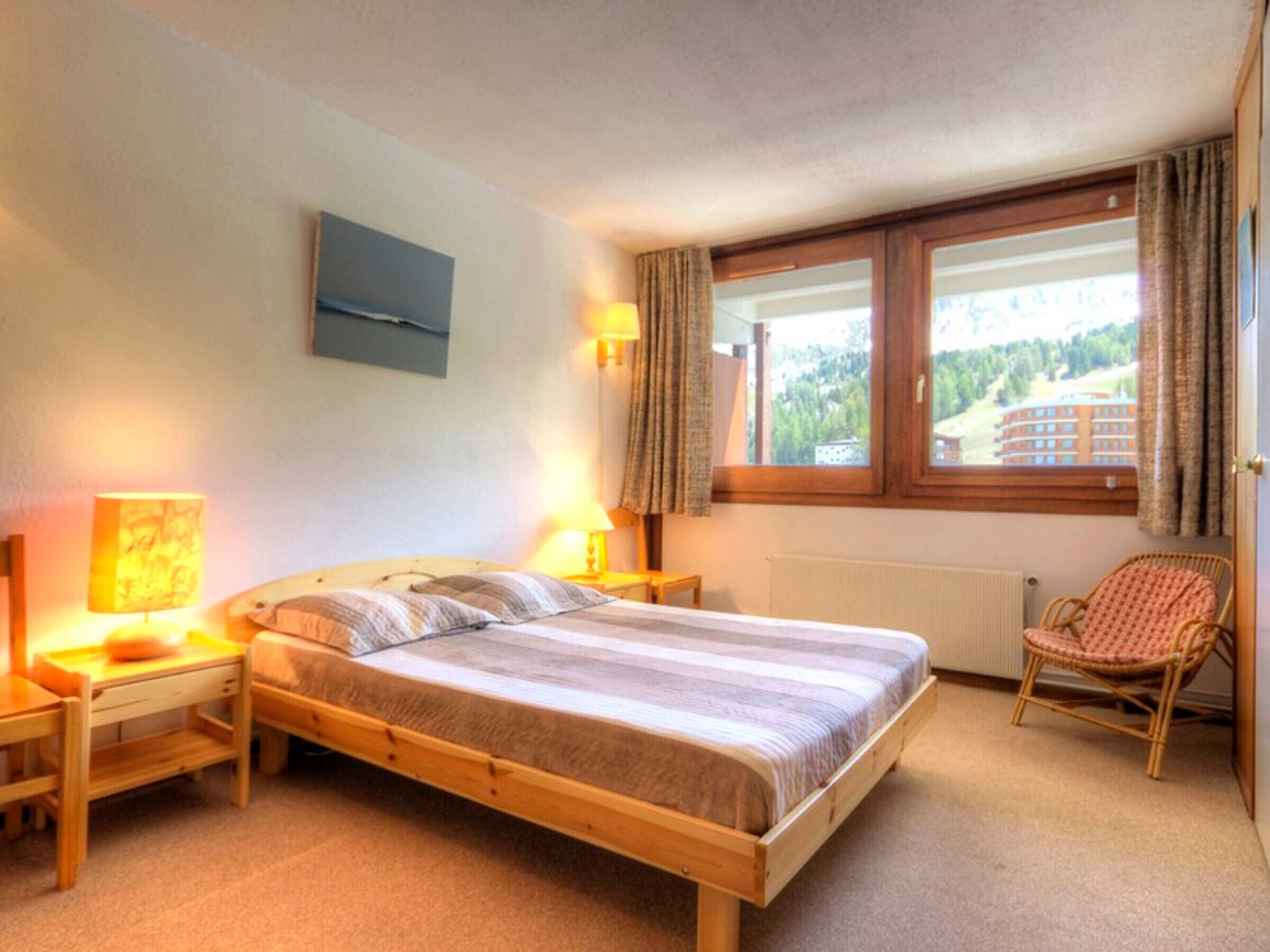 Een slaapkamer met een houten bed, twee bedlampen, een stoel, een raam met gordijnen en uitzicht op gebouwen.