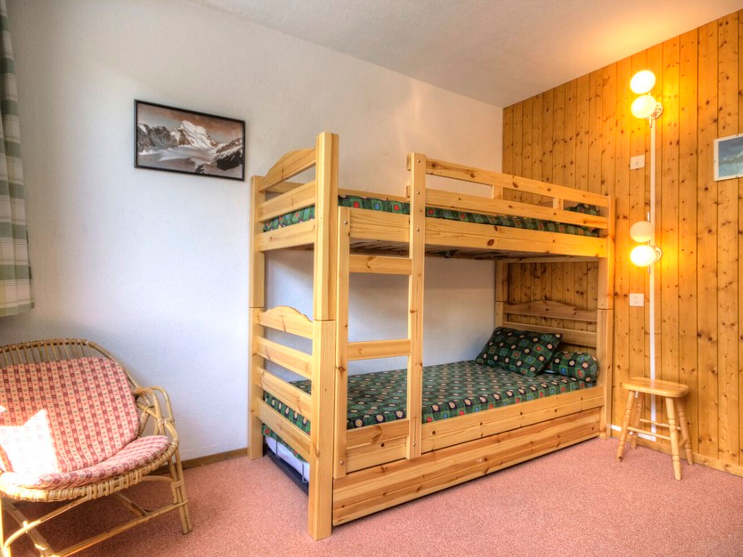 Un letto a castello con struttura in legno, un letto singolo con materasso a fantasia, una sedia in vimini e uno sgabello.