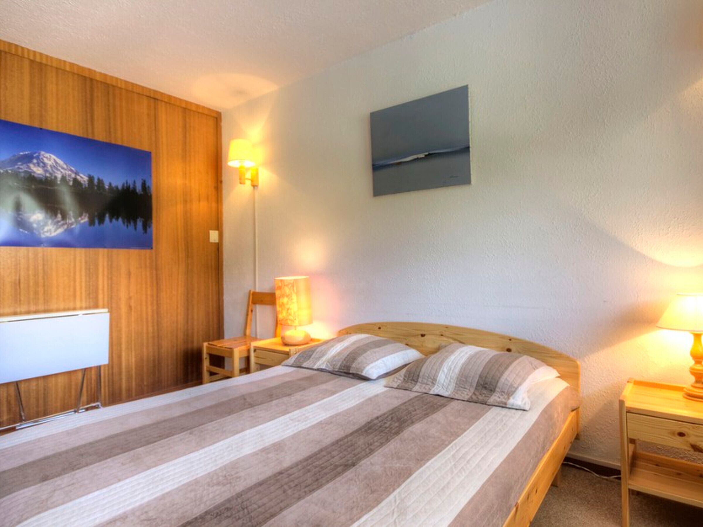 Ein Schlafzimmer mit Bett, Holz-Kopfstück, zwei Nachttischen mit Lampen, einem Sessel, einem an der Wand montierten Fernseher und zwei Bildern.