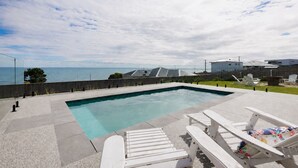 A rectangular pool with a clear blue edge, surrounded by a concrete deck. There are white lounge chairs with colorful cushions and a wooden fence in the background. - The Bay View San Remo by Ready Set Stay (San Remo)