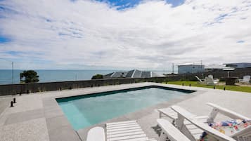 A rectangular pool with a clear blue edge, surrounded by a concrete deck. There are white lounge chairs with colorful cushions and a wooden fence in the background.