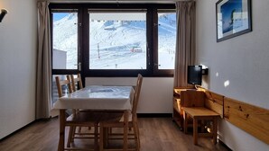 Una habitación con una mesa y sillas de madera, un escritorio pequeño de madera, un televisor y un ventanal con vistas a una montaña nevada.
