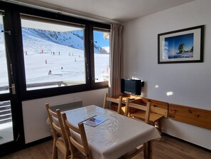 A room with a large window overlooking a snowy outdoor area with skiers. There is a wooden table with a white tablecloth, a television on a stand, and a framed picture on the wall.