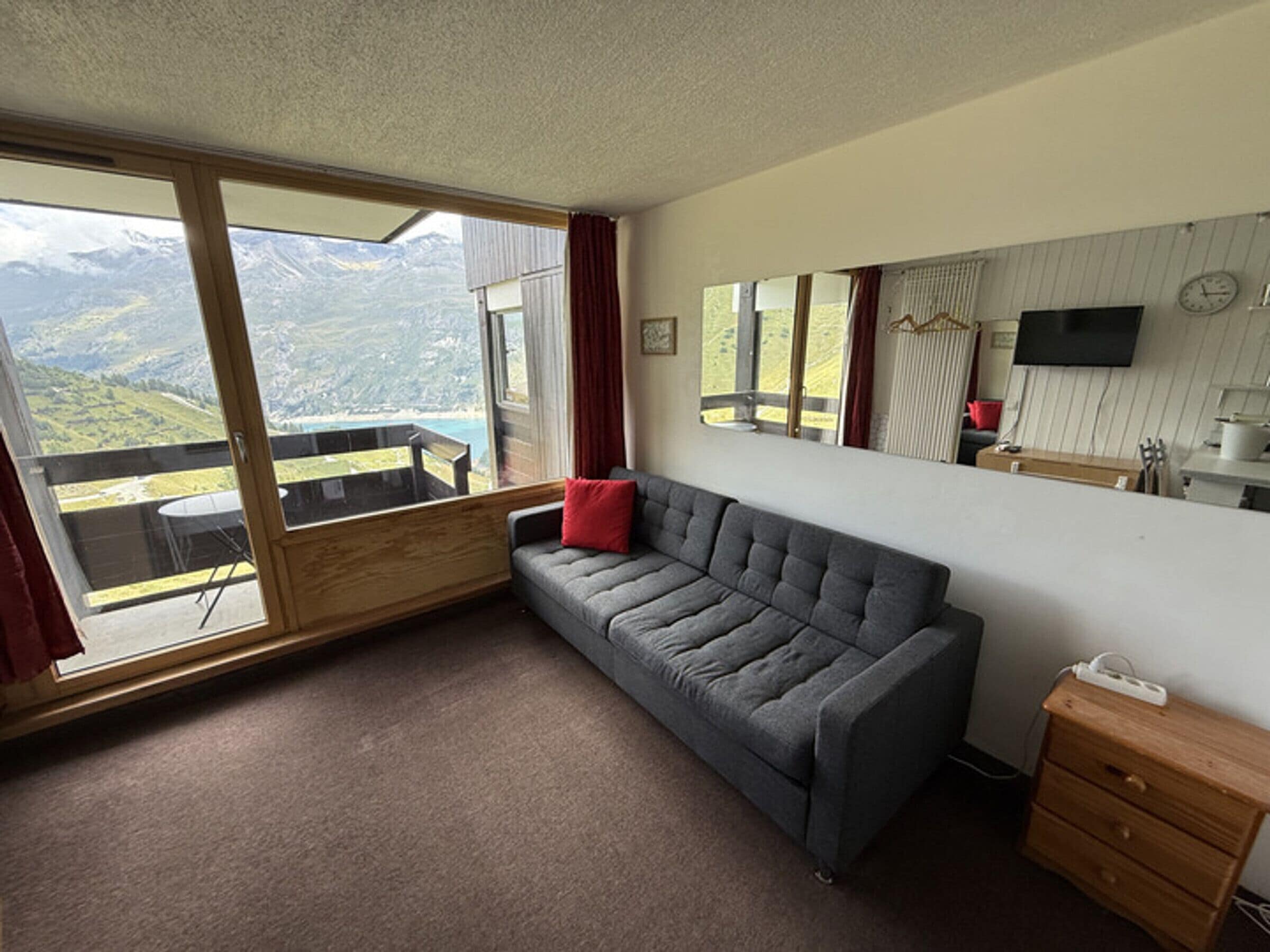 A room with a grey sofa, a wooden dresser, a large window with a mountain view, a mirror, and a flat-screen TV.
