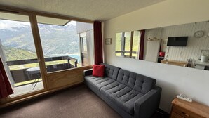 A room with a grey sofa, a wooden dresser, a large window with a mountain view, a mirror, and a flat-screen TV. - Comfortable studio with balcony, pets allowed, near slopes (Tignes)