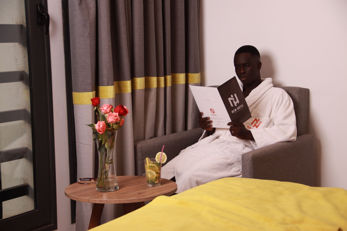A person sitting on a bed in a hotel room, reading a book. There is a vase with flowers on a side table.