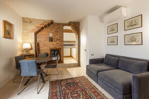 A living room with a grey sofa, a dining table with chairs, a patterned rug, and a brick wall with a clock.