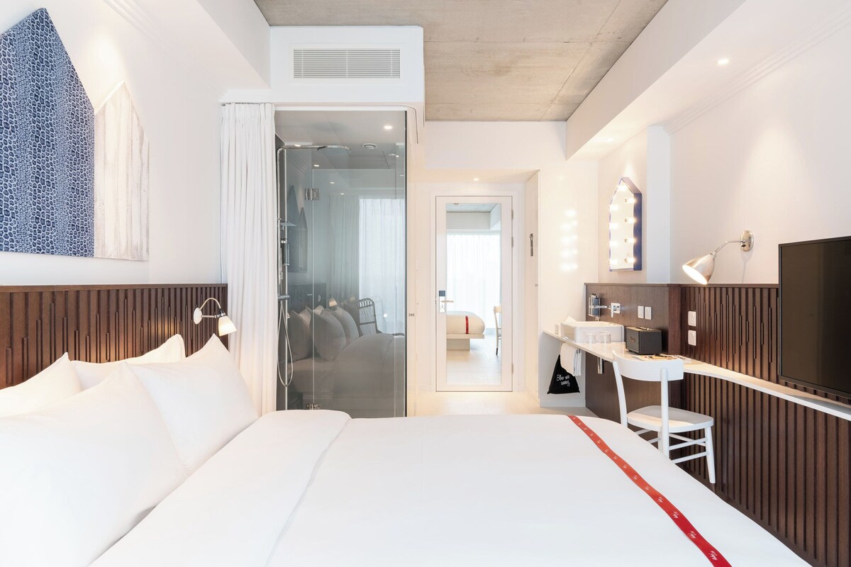 A hotel room with a bed, a vanity with a mirror, a television, and a shower area.