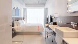 A modern hotel room with a large bed, a glass partition, a bathroom with a bathtub, and a large mirror with lights. - Ruby Zoe Hotel London by IHG (London)