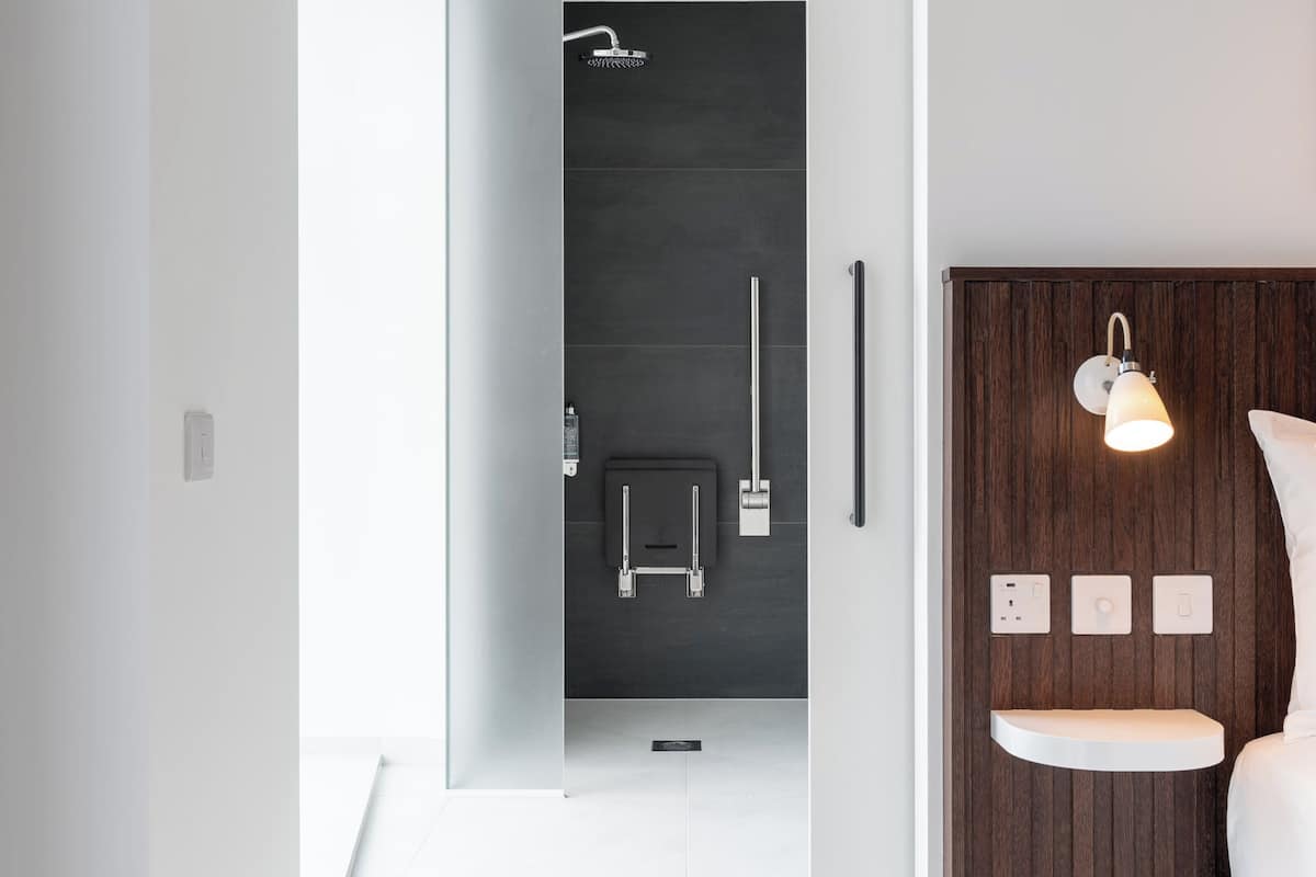 A modern bathroom with a shower, a toilet, and a sink. There is a wooden panel wall with a light fixture and a bedside table with a pillow.
