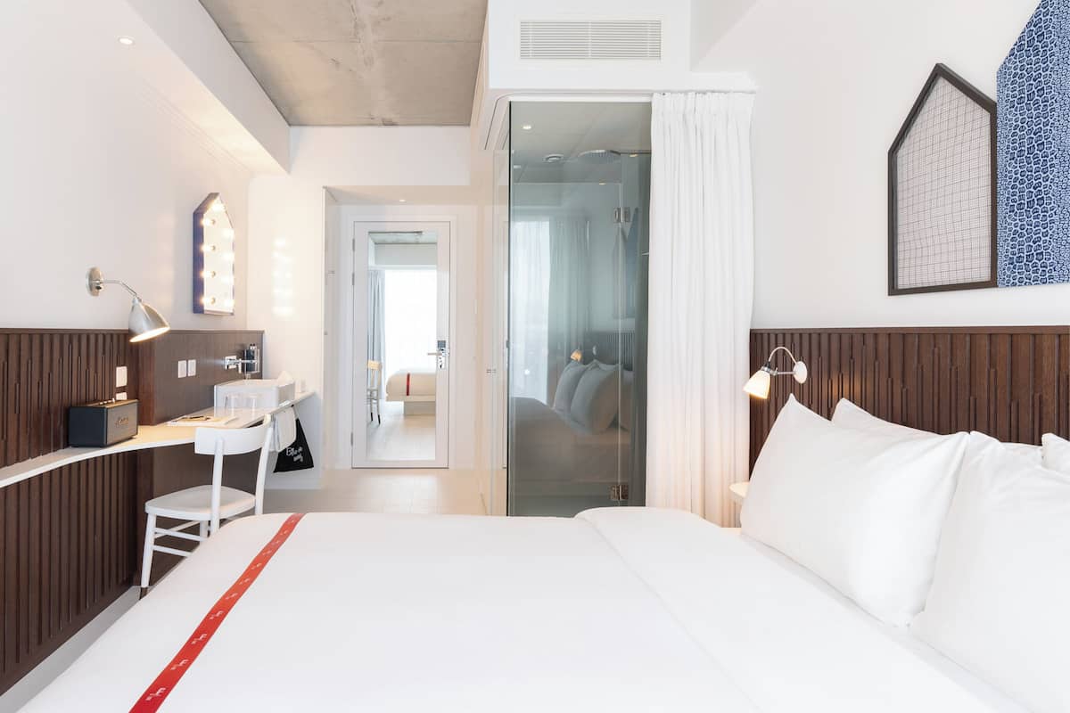 A hotel room with a large bed, a TV, a desk with a mirror, and a bathroom visible through a glass door.