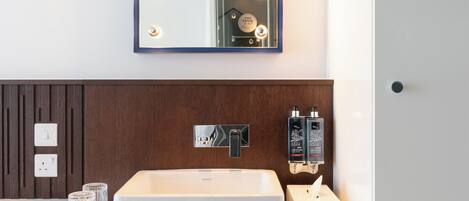 A modern bathroom with a large rectangular sink, a mirror with lights, a wooden panel wall, and two bottles of lotion.