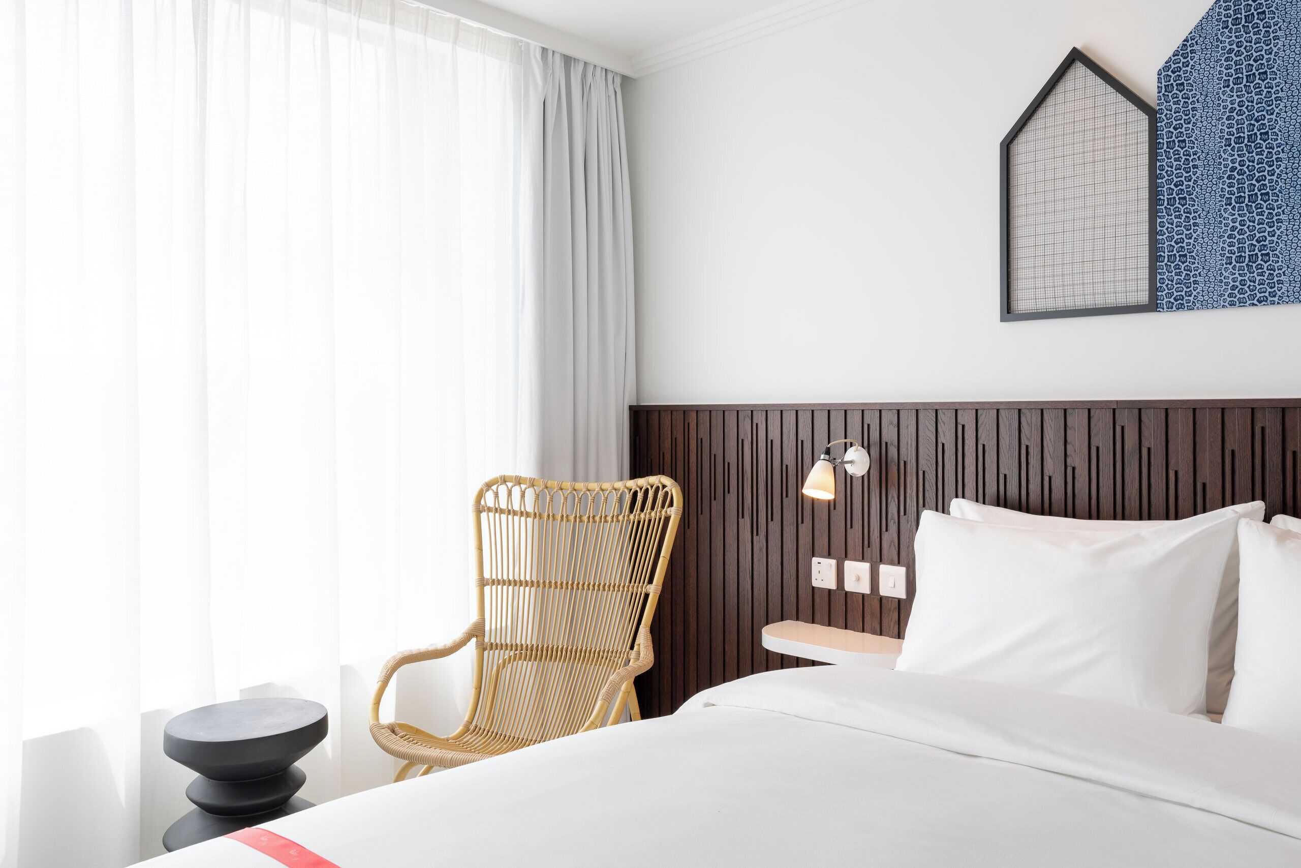A hotel room with a bed, a chair, a bedside table, and a wall-mounted lamp.