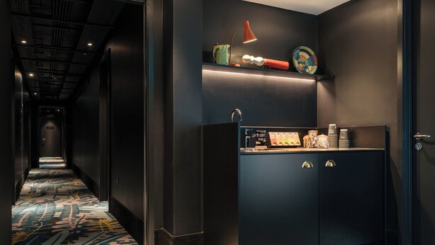 A hallway with patterned carpet, a dark wall, and a cabinet with a shelf above it. - Ruby Zoe Hotel London by IHG (London)