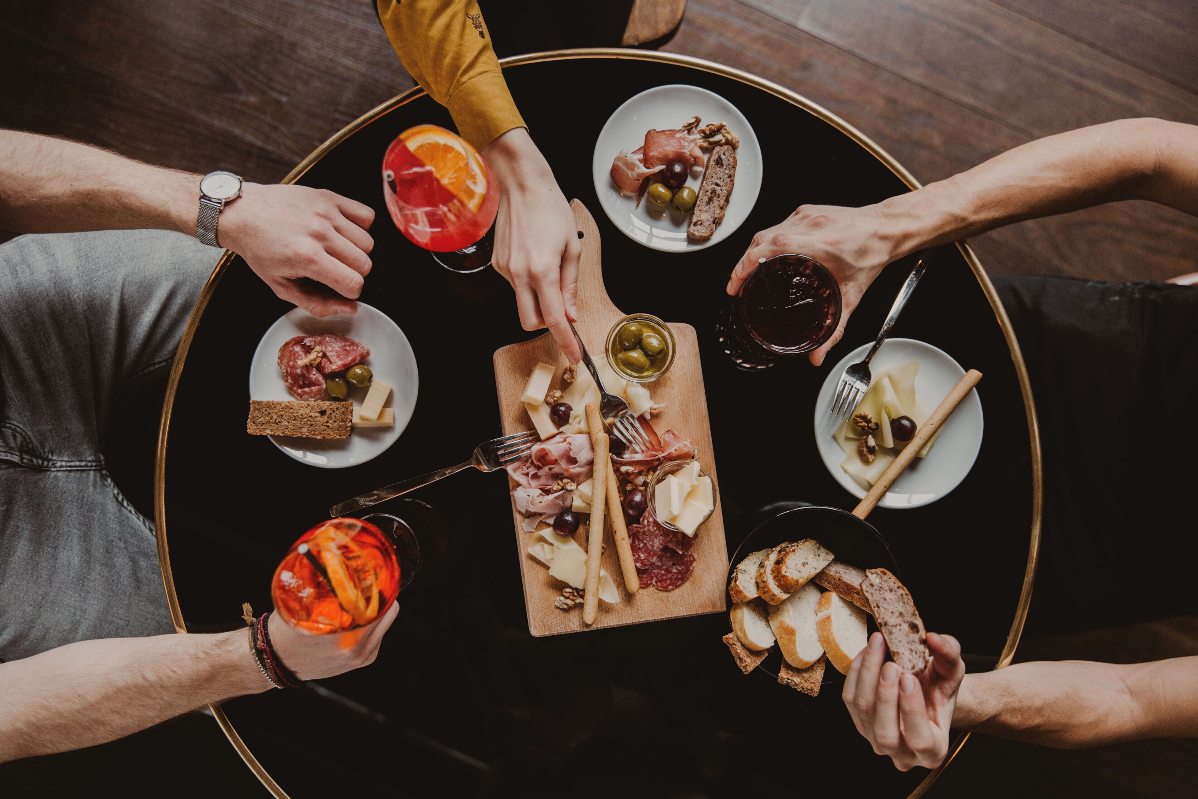 Una mesa redonda con diversos alimentos y bebidas, rodeada de gente.