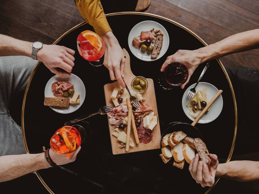 Una mesa redonda con diversos alimentos y bebidas, rodeada de gente.