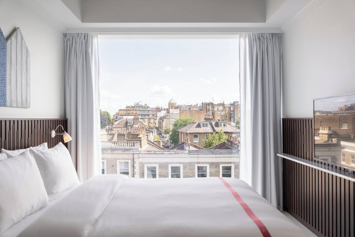 A hotel room with a large bed, a view of the city, and a TV on the right.