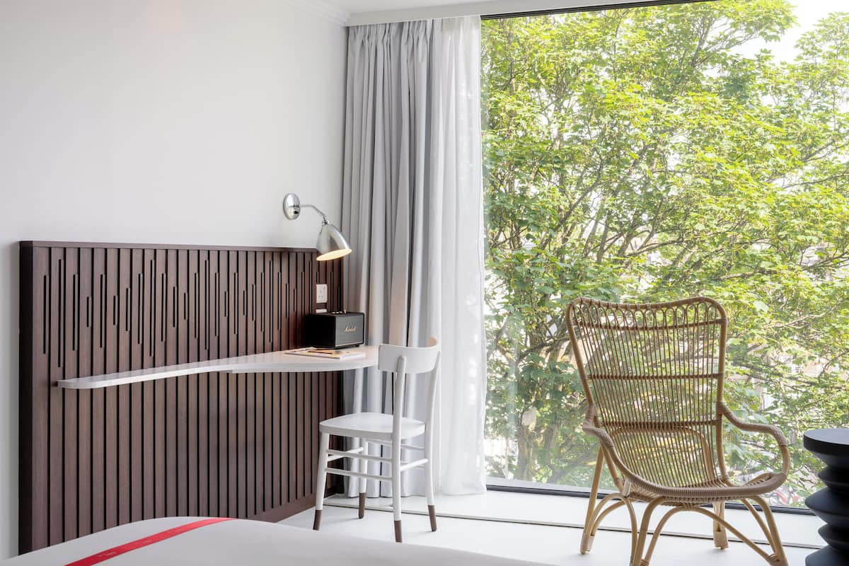 A modern room with a bed, a desk with a lamp, a wicker chair, and a large window with a view of trees.
