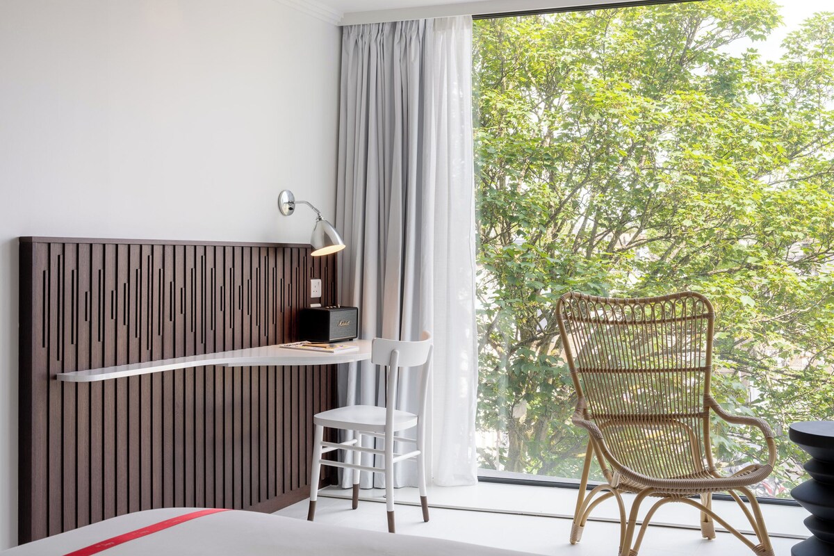 A modern room with a bed, a desk with a lamp, a wicker chair, and a large window with a view of trees.