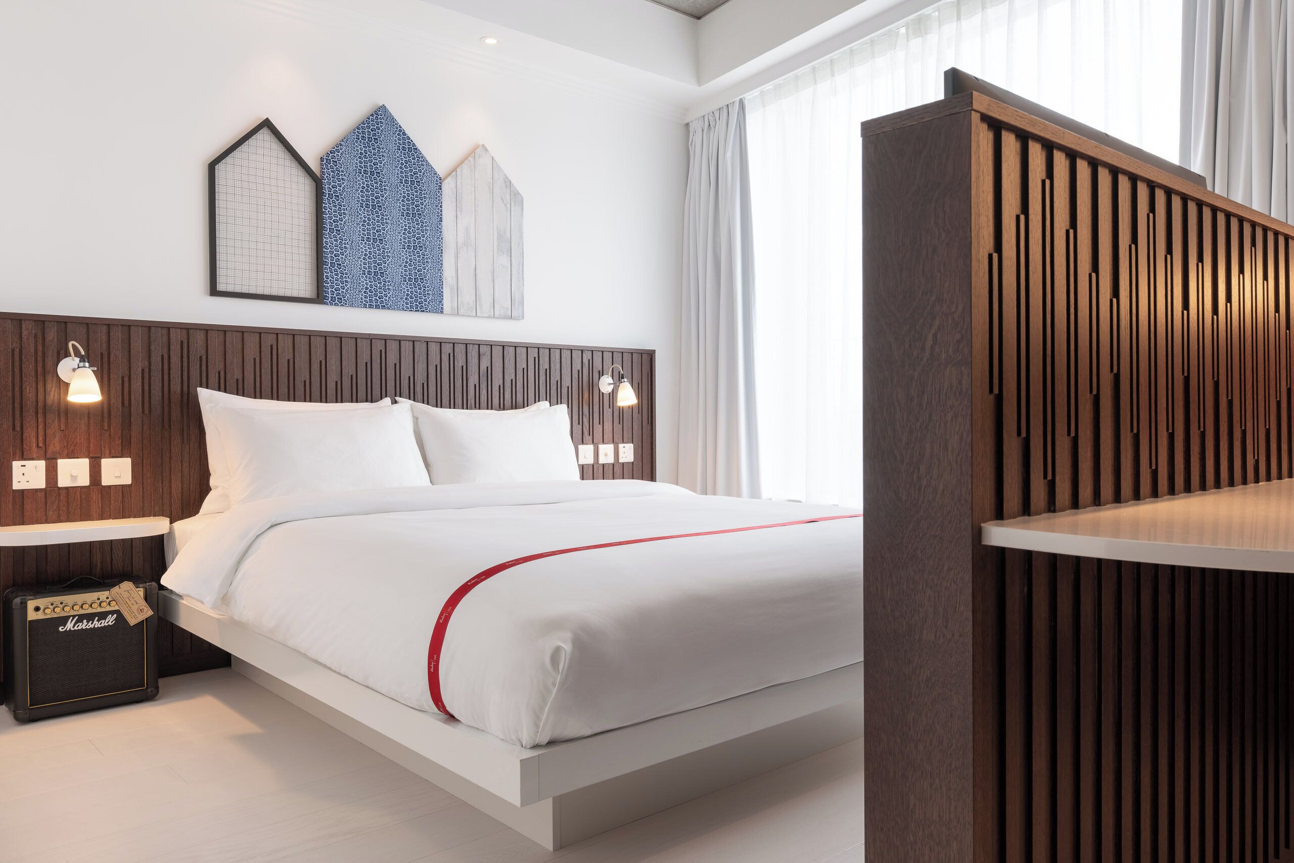 A modern hotel room with a wooden headboard, a white bed with red accents, a Marshall amplifier, and wall-mounted sconces.