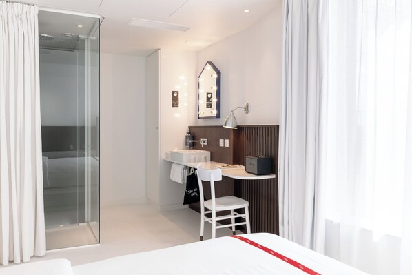 A modern hotel room with a glass shower enclosure, a vanity with a mirror, and a bed with a red accent.