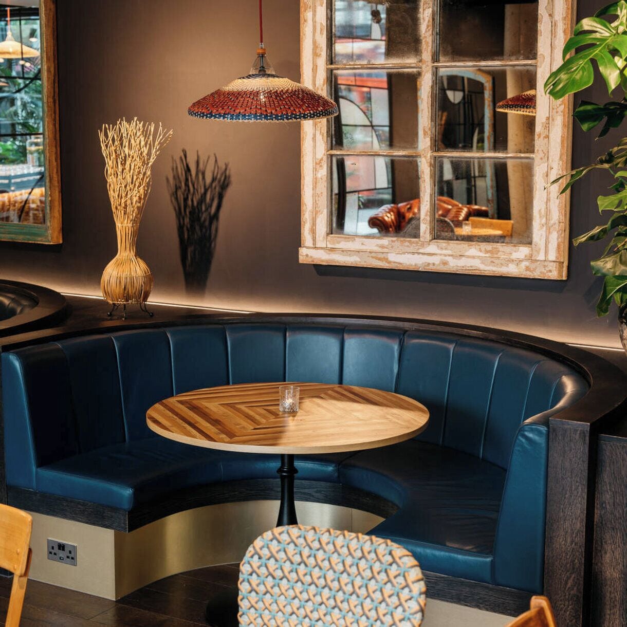 A curved blue bench with a round wooden table in the center, a woven lamp, and a window with a view of the street.