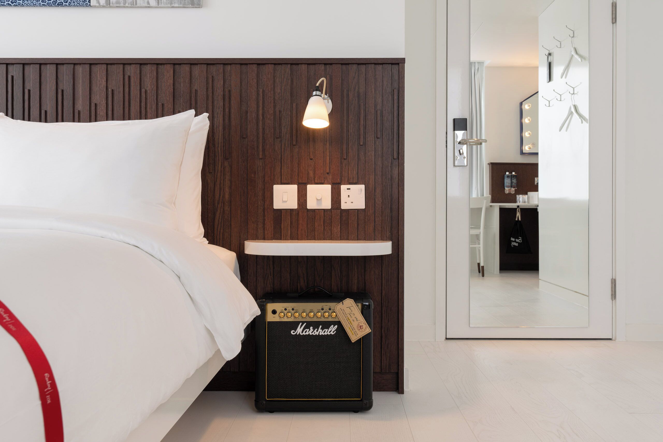 A bedroom with a bed, a Marshall amplifier, a wall-mounted light, and a mirror reflecting a bathroom.