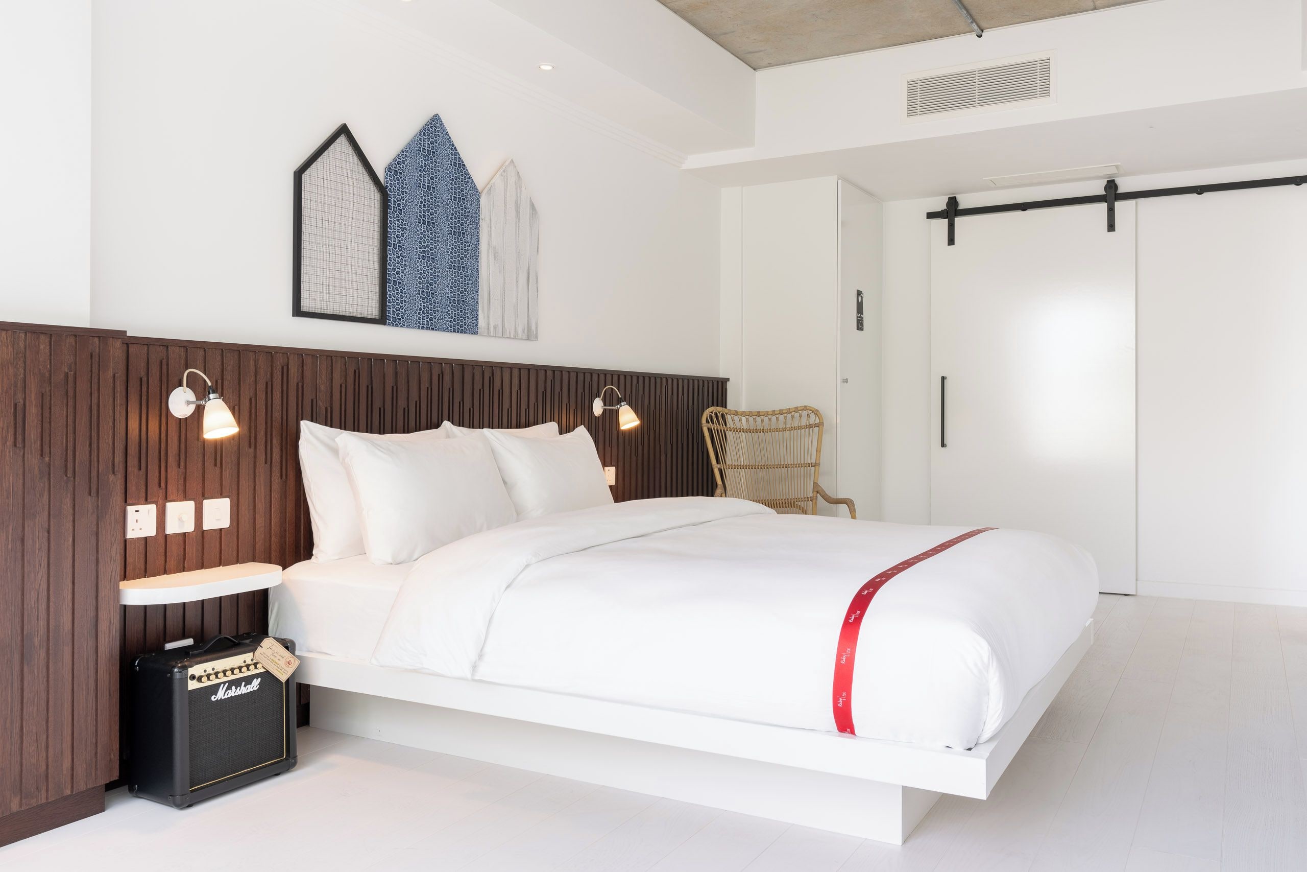 Un dormitorio moderno con cama, dos lámparas de noche, una obra de arte en la pared, cabecera de madera y un amplificador Marshall en el suelo.